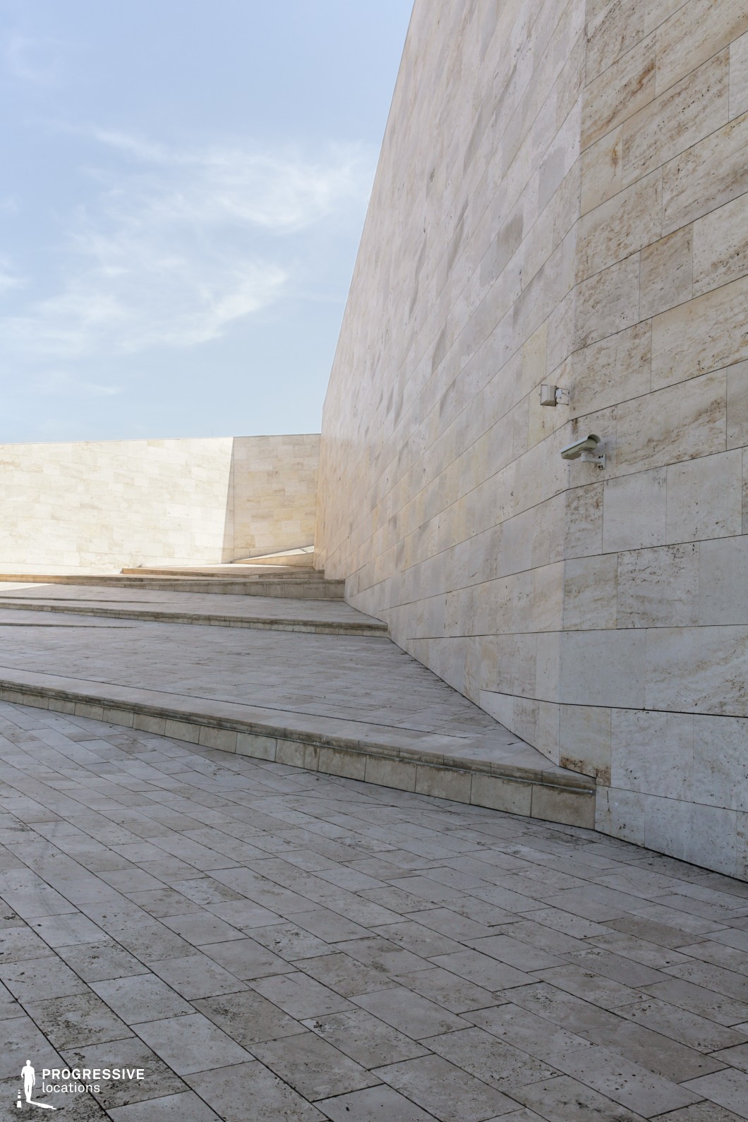 The image features a sleek, modern architectural structure with a curving stone staircase leading up to a tall, angular wall. The smooth, light-colored stone tiles cover both the floor and the wall, creating a harmonious, uniform surface that emphasizes the clean lines and geometric precision of the design. The gentle incline of the stairs contrasts with the towering, straight lines of the surrounding wall, producing a dynamic architectural scene. This location is ideal for filming contemporary urban scenes, architectural showcases, or films requiring minimalistic and stylish backdrops. Its angular shapes, smooth surfaces, and openness make it versatile for a range of film genres, including modern thrillers, dramas, or design-centric projects.