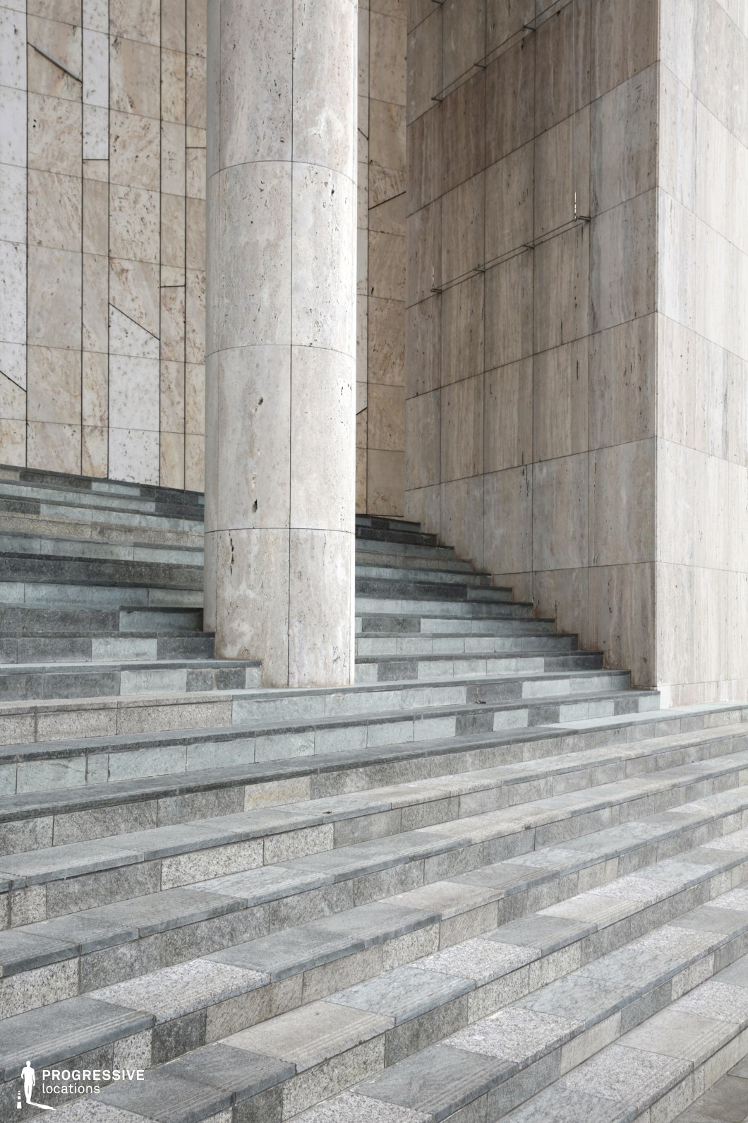 The image depicts a sophisticated stone staircase made of large, rectangular stone tiles, smoothly rising towards a prominent stone wall. The wall is textured with a contrasting finish, creating an elegant backdrop for the staircase. The steps are bordered by a column positioned to the side, further emphasizing the grandeur of the space. The polished stone textures and the sleek, linear design of the stairs provide a modern yet timeless feel, while the strong vertical column adds a sense of stability and monumentality. This location is perfect for filming scenes requiring formal or monumental backdrops, ideal for historical dramas, political thrillers, or architectural showcases, offering a balanced mix of modern and classical architectural elements.