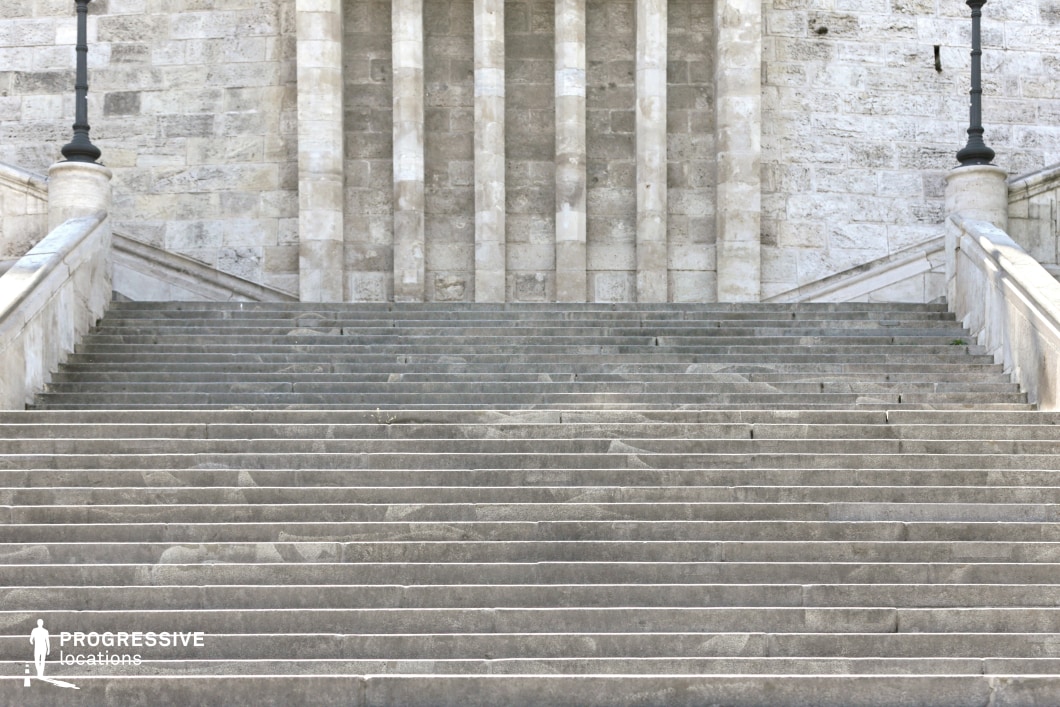 The image depicts an impressive stone staircase that leads up to a grand building, flanked by large columns. The staircase is made of solid stone, with clearly defined steps, each slightly worn with use, indicating the historical significance of the location. The columns rise behind the stairs, complementing the strength and formality of the structure. On either side of the stairs, traditional lamp posts add an elegant touch, further enhancing the monumental atmosphere. This location is ideal for filming grand, formal scenes, particularly in genres like historical dramas, period films, or action thrillers. The combination of the sturdy stonework, columns, and lamp posts creates a strong, dignified setting perfect for scenes of authority, power, or solemnity.