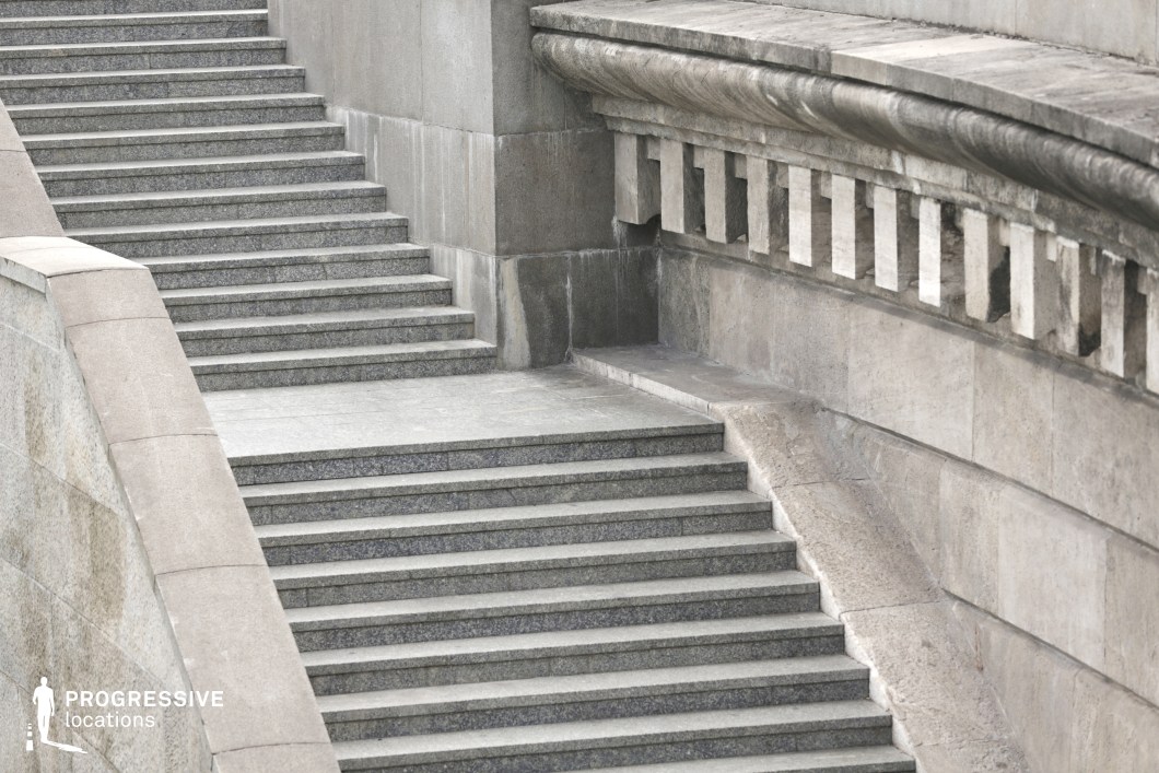 The image features a series of stone stairs, leading upwards with smooth, even steps that have a polished, modern appearance. The stairs are flanked by a decorative stone wall with a curved edge, adding an elegant architectural detail. The wall features rectangular cutouts and an intricate design along the top, contributing to a refined and structured atmosphere. The combination of the clean stone surfaces and the ornate detailing on the wall creates a sense of grandeur and timelessness. This location is ideal for filming scenes in historical dramas, formal gatherings, or films requiring a sophisticated, monumental backdrop. The strong visual lines and decorative elements provide a perfect setting for scenes of authority, transition, or ceremonial moments.