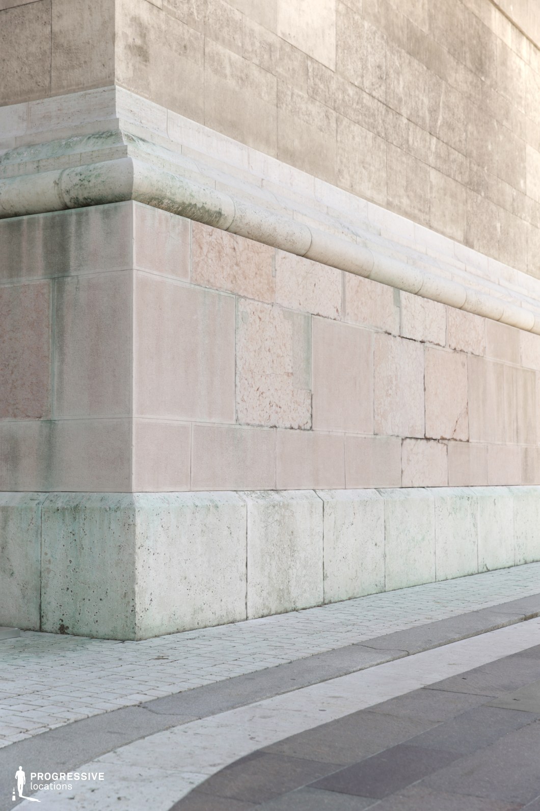The image presents a corner of a large stone wall, with smooth, weathered stone blocks in light shades of beige and pale gray. The base of the wall is finished with a clean, rounded stone molding, enhancing its architectural elegance. Below, a cobblestone path gently curves, complementing the aged stone texture and adding a sense of historical charm to the setting. The materials and textures of both the wall and the path convey a sense of timelessness and durability. This location is ideal for filming period dramas, historical scenes, or urban settings that require a blend of ancient and classic architectural styles. The visual appeal of the wall and the cobblestones creates a rich backdrop, perfect for films that highlight architecture, history, or the passage of time.