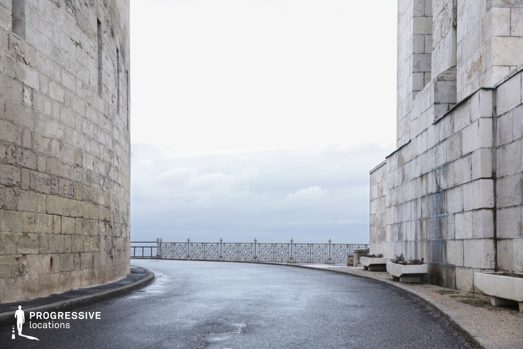 The image depicts a curved road lined by a stone wall on the left, leading to a panoramic view bordered by an ornate metal railing. The stone wall has a weathered, textured appearance, with large stone blocks adding a sense of strength and permanence. The road gently curves around the wall, offering a clear view of the horizon. The metal railing provides an elegant touch, contrasting with the rough stone. This location is perfect for filming scenes requiring a blend of natural beauty and architectural strength, making it ideal for dramatic or contemplative sequences, urban thrillers, or romantic films. The combination of the scenic curve, stone texture, and railing offers a versatile backdrop for various creative projects.