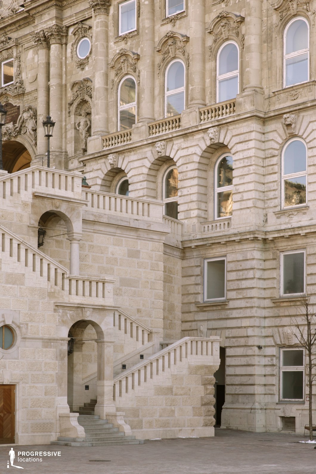 The image displays a grand staircase leading to a majestic building with intricately designed classical architecture. The staircase, made of light-colored stone, features elegant, curved arches and columns that highlight its monumental nature. The building’s facade is adorned with ornate window arches and decorative reliefs, contributing to the overall grandeur of the structure. The smooth texture of the stonework contrasts beautifully with the linear pattern of the stairs, which guide the viewer's eye upward. This location exudes a sense of opulence and historical richness, making it an excellent choice for filming scenes with regal or classical themes. The combination of architectural details and the dramatic staircase makes it ideal for historical dramas, fantasy films, or action scenes requiring a monumental setting.