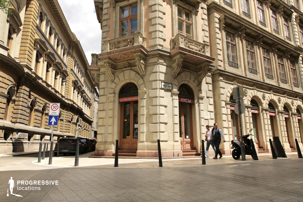 This image highlights a grand corner building with classical architectural details, featuring large windows, intricate stonework, and a prominent balcony above the main entrance. The cobblestone street in front of the building complements the historic vibe, while the well-maintained facade showcases the building's classical elegance. The clear lines of the building, combined with the detailed stone textures, create a striking visual, ideal for portraying the opulence of a past era. This location is perfect for filming historical or period dramas, urban thrillers, or even romantic scenes, offering a blend of old-world charm and dynamic urban energy. The corner positioning of the building and the detailed streetscape make it suitable for establishing shots, exterior interactions, or sequences requiring a stately, timeless atmosphere.