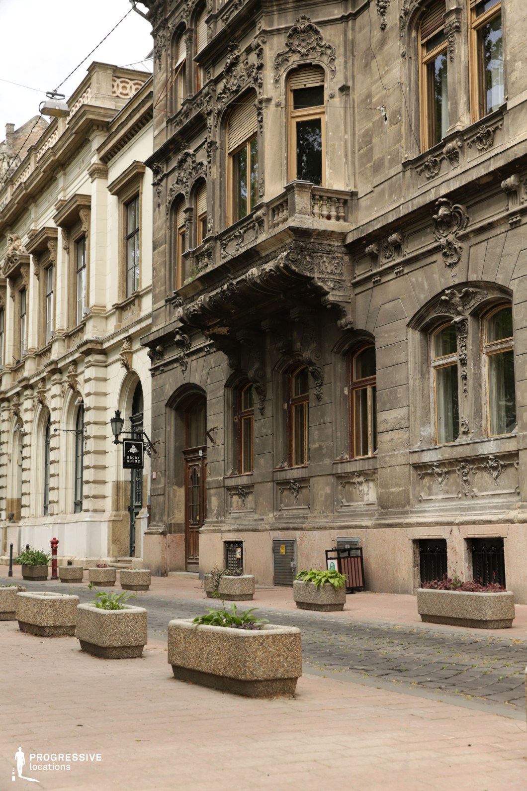 The image features a distinguished architectural facade with a blend of classical and intricate detailing. The building’s stonework showcases elaborate window frames, decorative carvings, and ornamental balconies, reflecting a sense of grandeur and historical significance. The cobblestone pavement in front adds a rustic touch, complementing the stately appearance of the building. The surrounding street is lined with simple planters, providing a touch of greenery to the otherwise formal and elegant setting. This location offers a timeless backdrop, perfect for films with historical, dramatic, or romantic themes. Its combination of classical architecture and cobblestone streets makes it ideal for capturing intimate moments or scenes set in urban environments of the past.