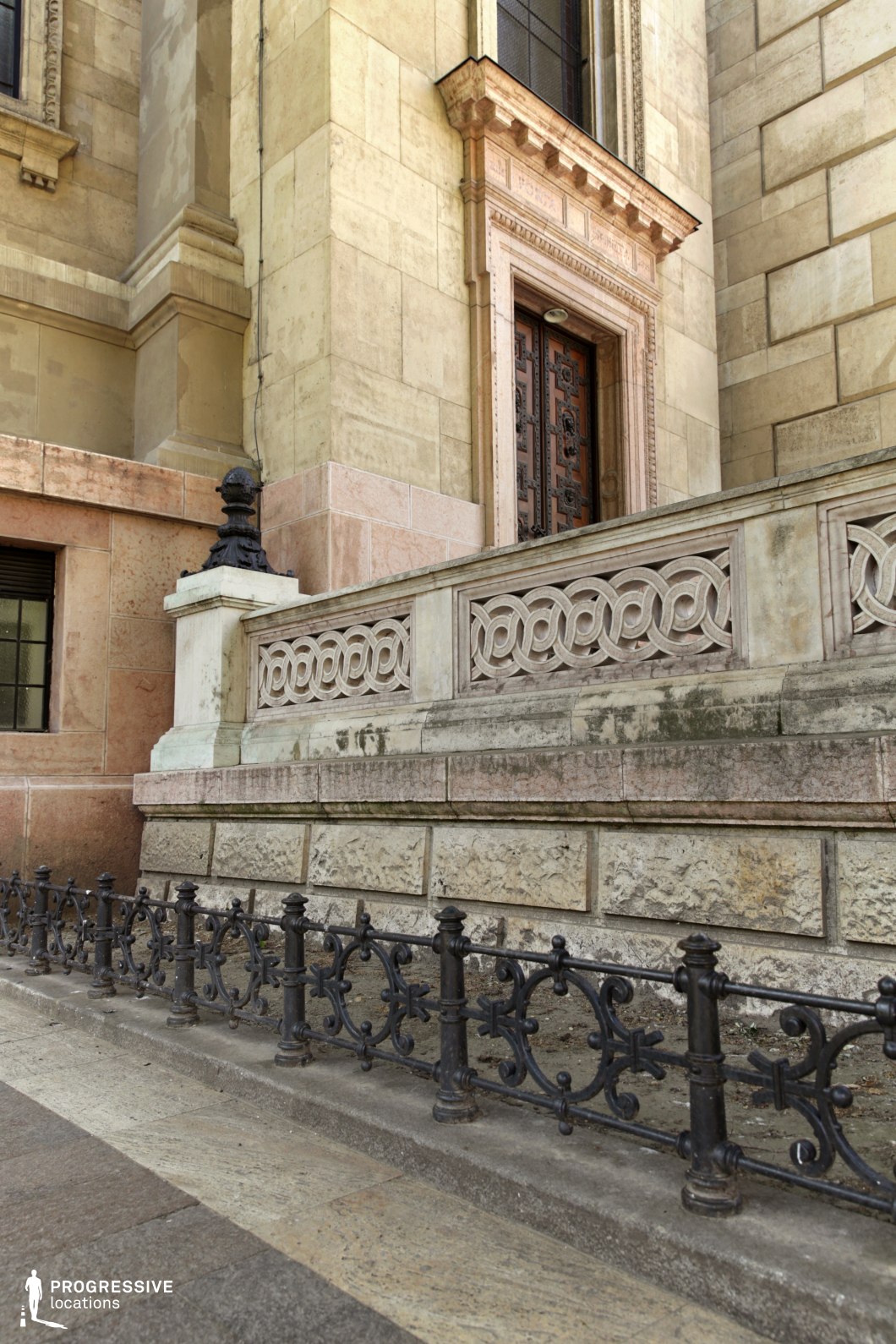 This image presents a detailed view of a grand building’s base, showcasing its intricate wrought iron fence and marble and limestone facade. The ornate fence runs along the stone path, adding a touch of elegance with its delicate, swirling patterns. The weathered stone pillars and the polished marble elements of the building reflect a classical style, with the door's detailed carvings standing out as a focal point. The combination of textures—smooth marble, rough limestone, and intricate wrought iron—creates a refined, historical atmosphere. This location is ideal for filming scenes requiring an air of sophistication and timelessness, suitable for genres such as historical dramas, romantic films, or detective mysteries. The elegant architectural details and sturdy design make it perfect for exterior shots, establishing scenes, or moments requiring a backdrop of grandeur and class.