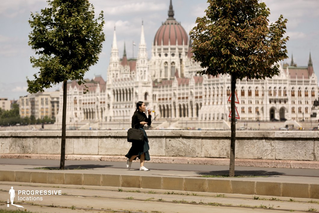 This image captures a scenic view of the Hungarian Parliament building, with its majestic, neo-Gothic architecture standing proudly in the background. The foreground features a clean, modern riverside pathway with trees lining the way, adding a touch of greenery to the urban landscape. The symmetry of the building's spires and its ornate detailing contrasts beautifully with the natural elements. The combination of the iconic landmark and tranquil riverside setting makes this an ideal location for urban or historical dramas. It offers a timeless backdrop for establishing shots, peaceful walks, or dramatic scenes requiring grandeur, making it perfect for political thrillers or romantic films in city settings.