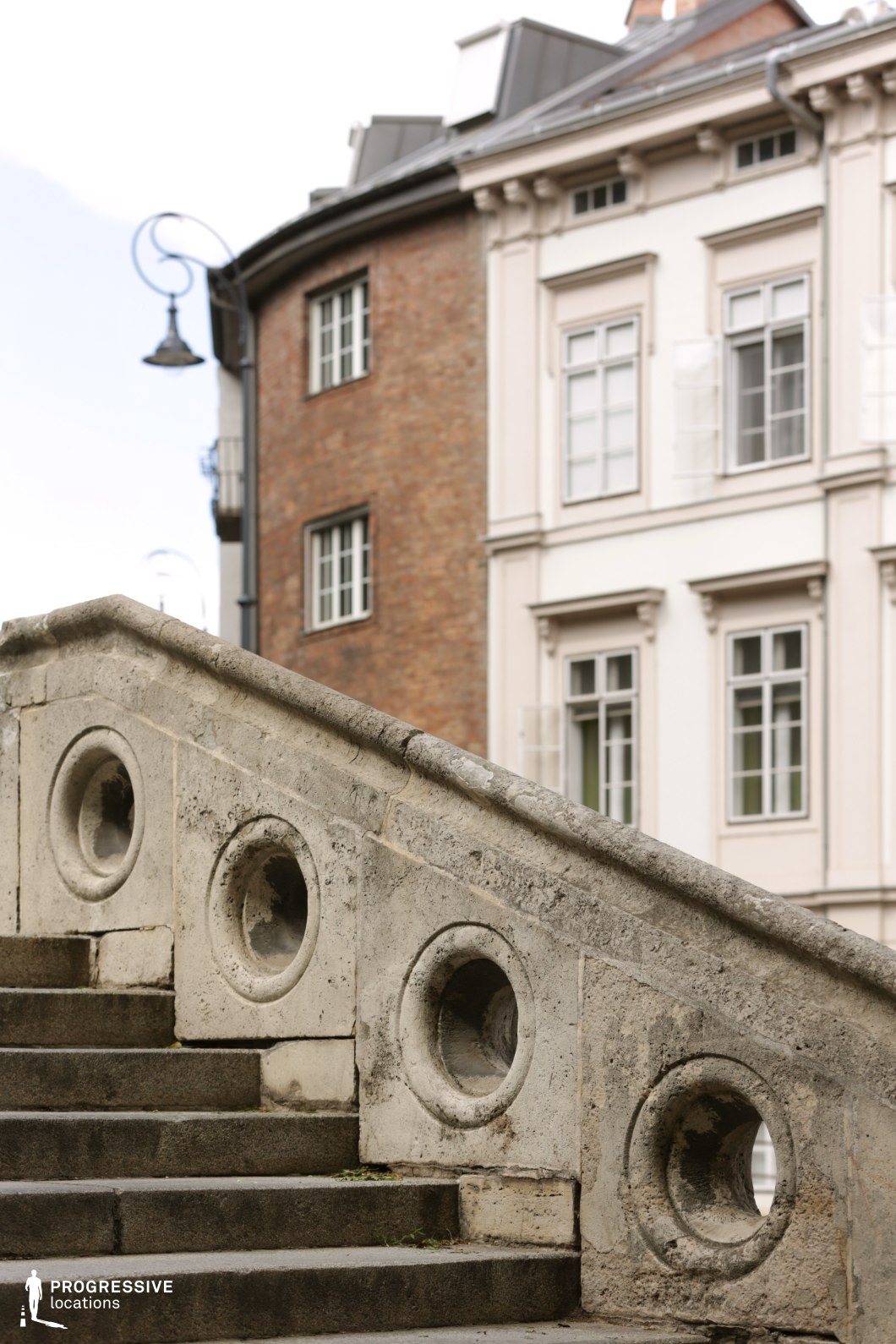 The image showcases a set of stone stairs with a unique decorative design, featuring large circular indentations along the railing. The stairs lead toward a charming Victorian-style building with detailed window frames and a smooth stone facade. The architectural contrast between the textured, weathered stairs and the sleek, well-maintained building emphasizes a blend of classic and more contemporary design elements. This location is ideal for films seeking a vintage or elegant urban backdrop, perfect for period dramas, romantic films, or historical narratives. The decorative elements and the architectural contrast between the stairs and the building create a visually rich setting for cinematic scenes.