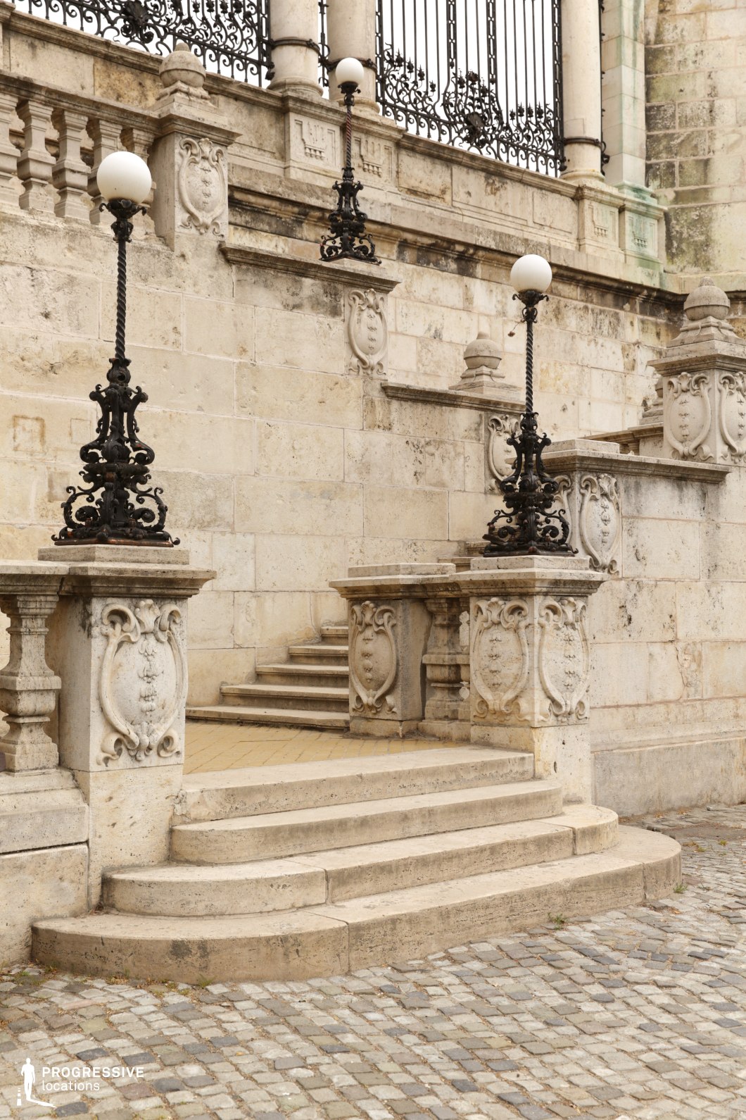 This image features a set of grand limestone stairs, elegantly flanked by ornate wrought iron lamp posts. The carved stone balustrades and decorative features add to the grandeur of the staircase, while the white spherical lights atop the lamps provide a vintage touch. The weathered stone surface and the cobblestone ground beneath suggest a long history, with the architecture showcasing fine craftsmanship. This location is perfect for filming scenes that require a majestic, classical atmosphere. It can be used in historical films, period dramas, or scenes with a sense of grandeur and elegance. The dramatic combination of intricate detailing and expansive stairs makes it ideal for establishing shots, formal entrances, or cinematic moments requiring a refined, timeless backdrop.
