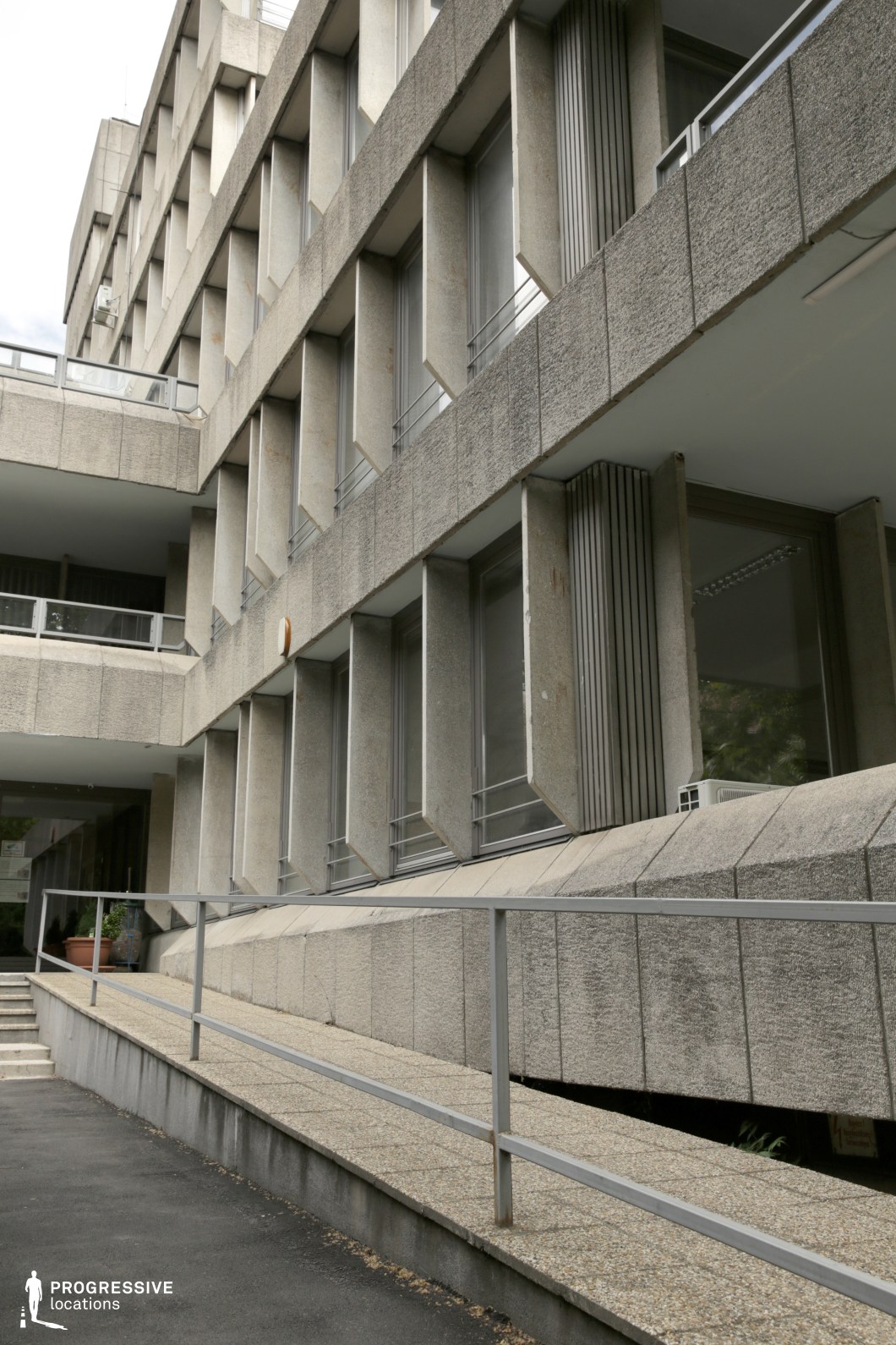 This modern concrete building features angular design elements, with textured concrete panels and rows of large windows framed by overhanging concrete. A metal railing runs along a ramp leading to the entrance, enhancing the building's industrial, minimalist aesthetic. The sharp lines and geometric patterns create a clean, functional appearance. This location is perfect for contemporary dramas, corporate settings, or films with futuristic themes, offering a sleek backdrop for urban or architectural-focused scenes.