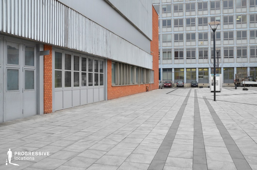 This location showcases a modern urban setting, with a blend of industrial and functional design elements. The foreground features a large, paved plaza with grey tiles, neatly arranged to create a grid pattern. The space is flanked by two contrasting building facades; one is a smooth, metallic grey with large windows, and the other features warm, textured brick walls with similarly spaced windows. In the background, a plain, boxy office building stands with a simple, grid-like window design. The surrounding space includes a few cars parked along the edge and modern street lamps. This location is ideal for filming in settings that require a clean, functional urban environment, suited for genres such as crime, drama, or corporate thrillers. The combination of sleek modern architecture and industrial elements offers a versatile backdrop for both dramatic and everyday city scenes.