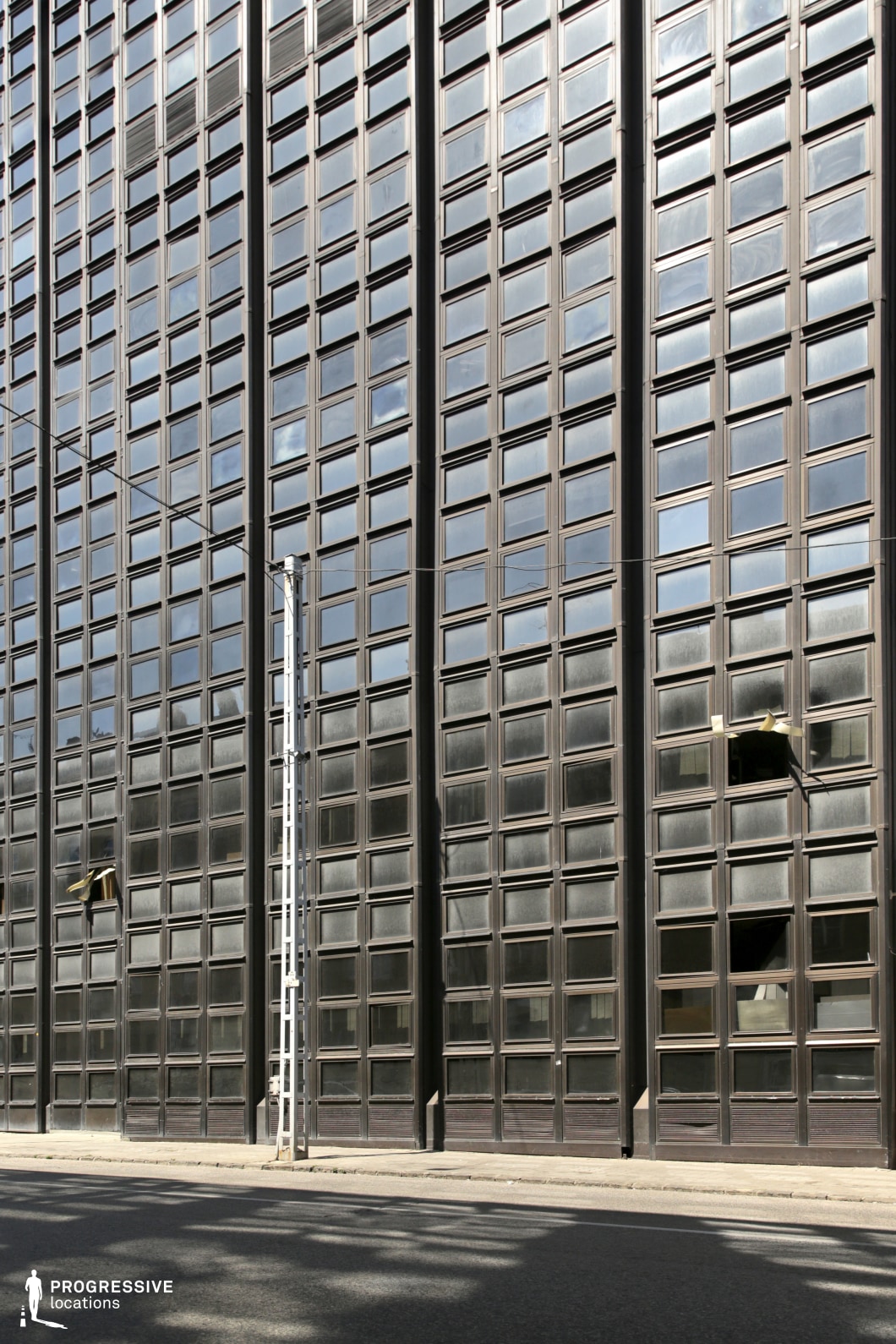 This image features a large, imposing high-rise building with a grid pattern of rectangular windows. The structure is composed of dark glass framed by metal, giving it an industrial and modern aesthetic. In front of the building, a narrow pavement runs along the base of the facade, with a metal ladder against the wall, adding a functional, utilitarian element to the scene. The building’s windows reflect the surrounding environment, contributing to the stark, geometric appearance. This location is ideal for filming in urban or corporate settings, making it suitable for genres like drama, thriller, or action. The symmetrical, grid-based design allows for striking cinematic compositions, emphasizing modern cityscapes, large-scale architecture, or corporate environments. The minimalistic features and clean lines also lend themselves well to scenes of high tension or sleek, futuristic settings.