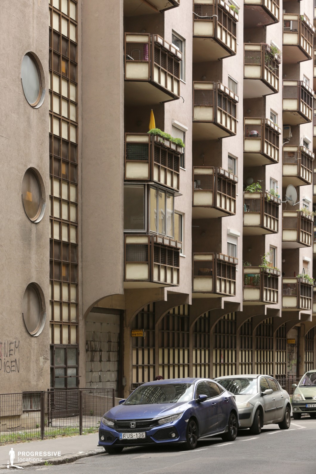 This residential building features protruding metal-framed balconies with plants, alongside rectangular and circular grid-patterned windows. The beige exterior gives it an industrial feel, while archways beneath the balconies add unique structural elements. Parked cars along the street highlight its urban setting. This location is ideal for urban dramas, architectural films, or scenes depicting city life, offering a backdrop of mid-20th-century residential design perfect for moments of tension or everyday interactions.