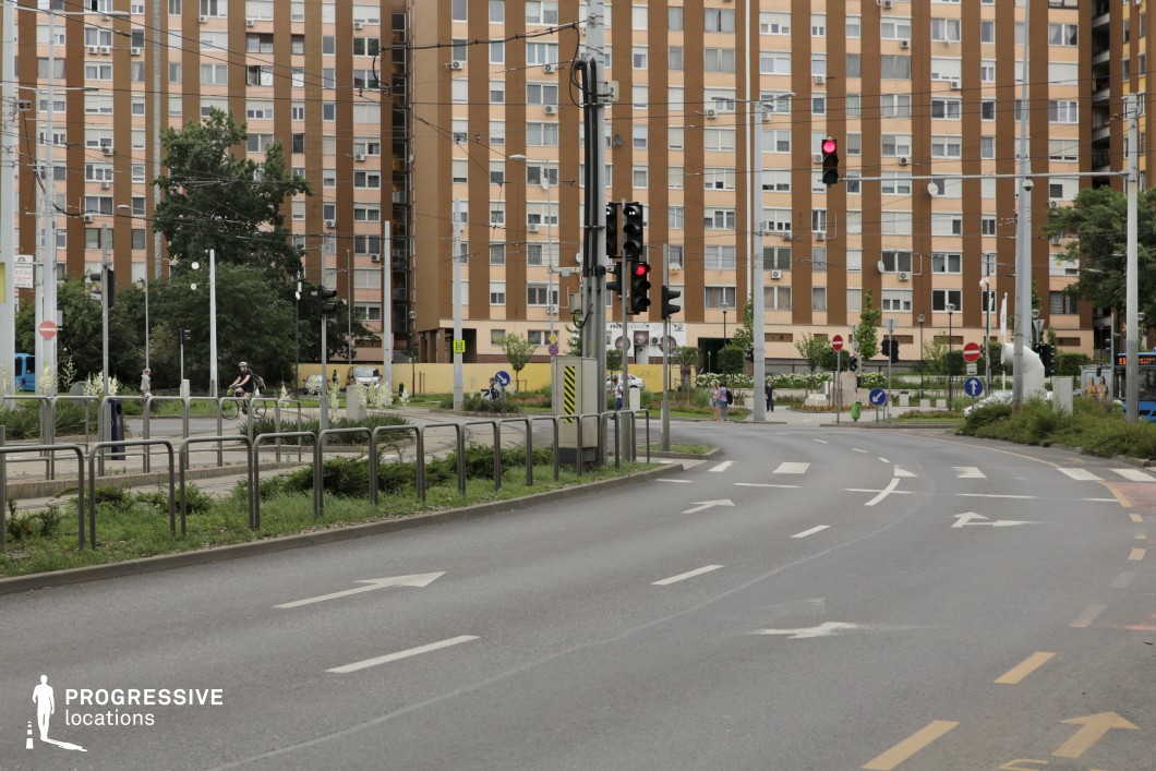 This urban street features clear road markings, traffic signs, and red traffic lights, with a prominent housing estate in the background, characterized by its brown facade and rows of windows. The street is lined with plants and trees, and cyclists use the bike lane, adding to the lively, functional atmosphere. This location is perfect for urban dramas, action scenes, or films set in a modern city, offering a realistic backdrop for scenes involving city life, transportation, or everyday urban interactions.
