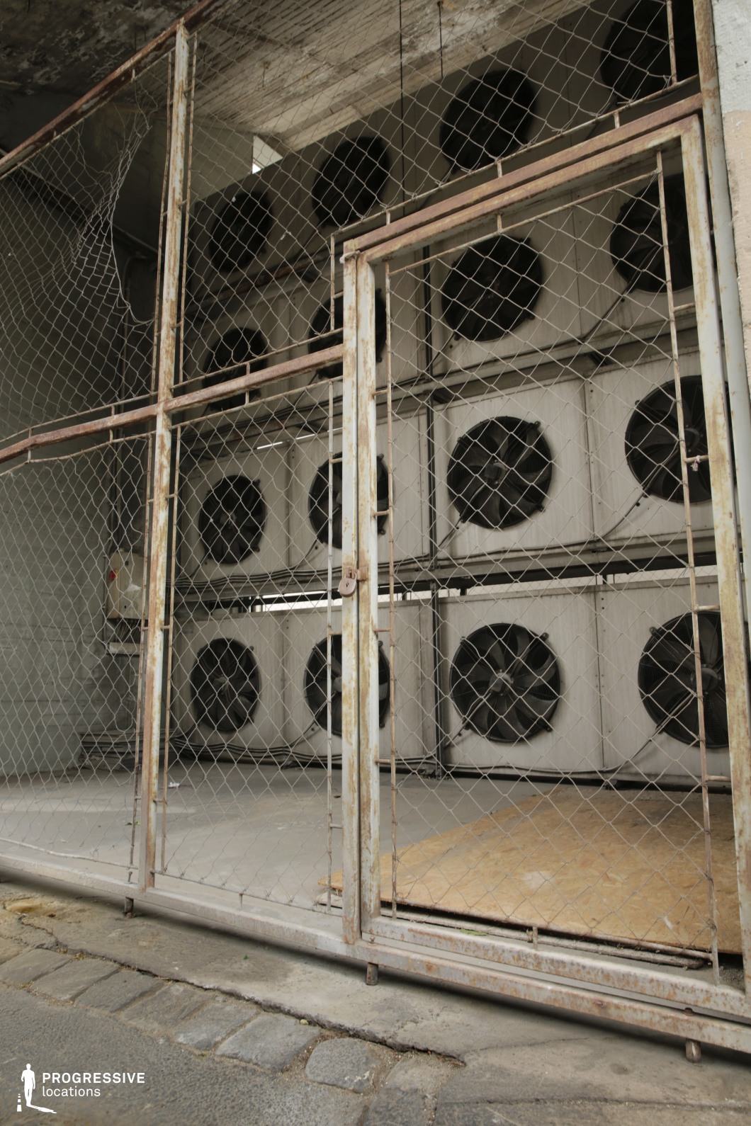 This image features large industrial cooling units enclosed by a rusted, chain-link fence, with visible fans behind each grate. The weathered fence and concrete floor enhance the gritty, utilitarian feel of the scene, emphasizing its industrial atmosphere. This location is perfect for industrial-themed films, dystopian settings, or thriller and crime scenes, offering a raw, factory-like backdrop that conveys urban decay and the inner workings of industrial areas.