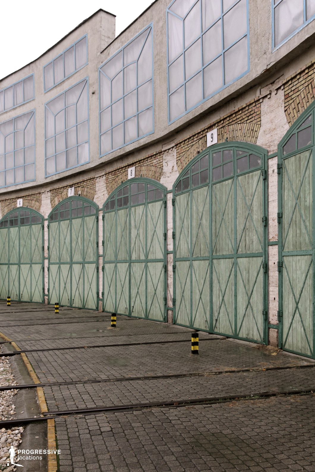 This railway depot features a series of green wooden gates framed by brick arches and numbered, with large rectangular windows above them. The cobblestone ground is lined with yellow-and-black striped bollards, and tracks curve along the edge of the depot. The weathered appearance of the gates and the industrial design evoke a sense of utilitarian efficiency. This location is ideal for industrial, historical, or action-themed films, offering a gritty backdrop for scenes set in working-class environments or post-apocalyptic narratives, with its distinctive architecture adding to the atmosphere of urban decay.