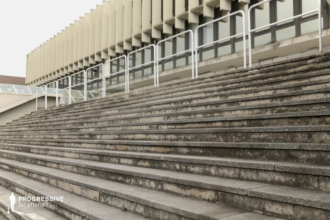 This architectural scene focuses on a set of wide, worn concrete steps leading up to a building with a distinctive modernist design. The building’s facade is made up of repeating grid-like windows, framed by a white railing that runs along the top of the staircase, providing both functionality and a clean aesthetic contrast. The worn texture of the concrete steps adds a sense of age and use, complementing the sleekness of the building’s windows. The building’s design, with its functional geometry and industrial appeal, is perfect for urban-themed films, particularly those in action, drama, or suspense genres. This location can effectively portray institutional or public spaces, with the stark concrete and steel details enhancing scenes of city life, modern architecture, or architectural decay.