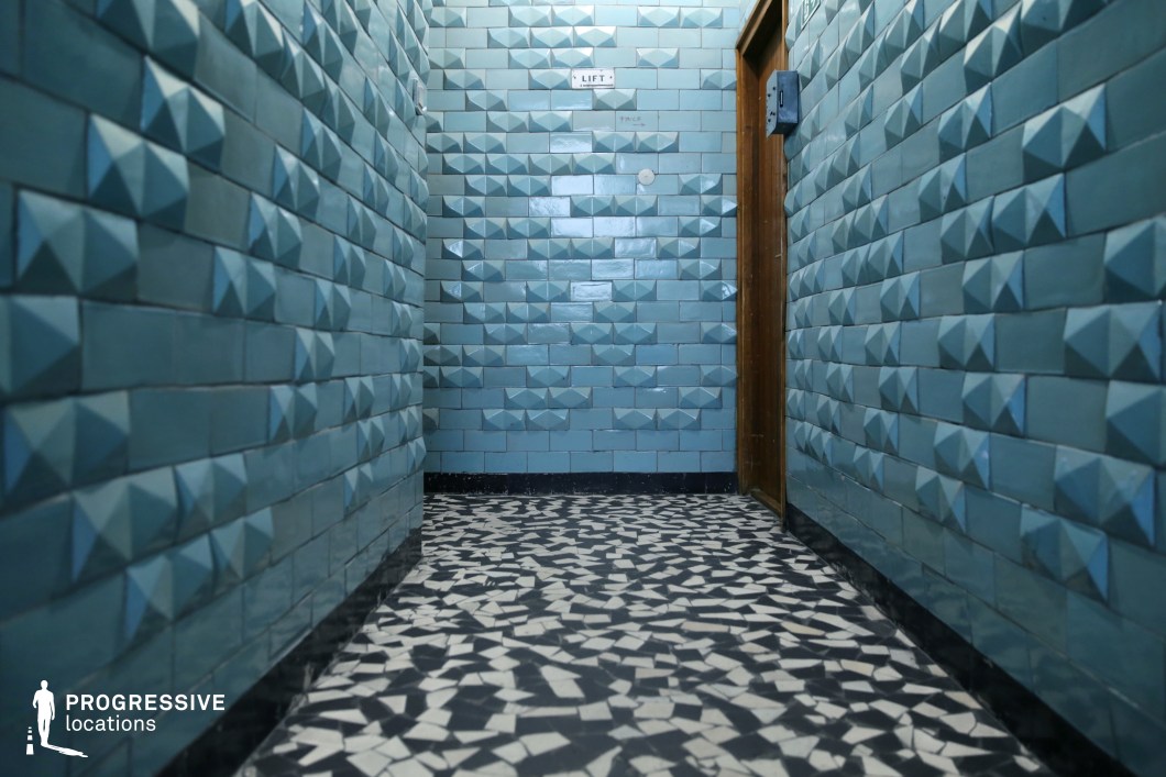 The image showcases a narrow corridor lined with distinctive blue tiles that have a pyramidal texture, creating a unique visual pattern. The walls feature a combination of matte and glossy tiles, adding depth and a subtle shine. The floor is covered with black and white mosaic tiles, contributing to the contrast and enhancing the geometric feel of the space. A wooden door with a simple design can be seen at the end of the corridor, alongside a sign indicating a lift. This location, with its modern and geometric aesthetics, is well-suited for film genres that require an edgy or industrial atmosphere, such as thrillers, mysteries, or science fiction. The bold color contrasts and unique tile designs would complement scenes in settings like hotels, office buildings, or futuristic environments, providing a visually striking backdrop for both tense and atmospheric moments.