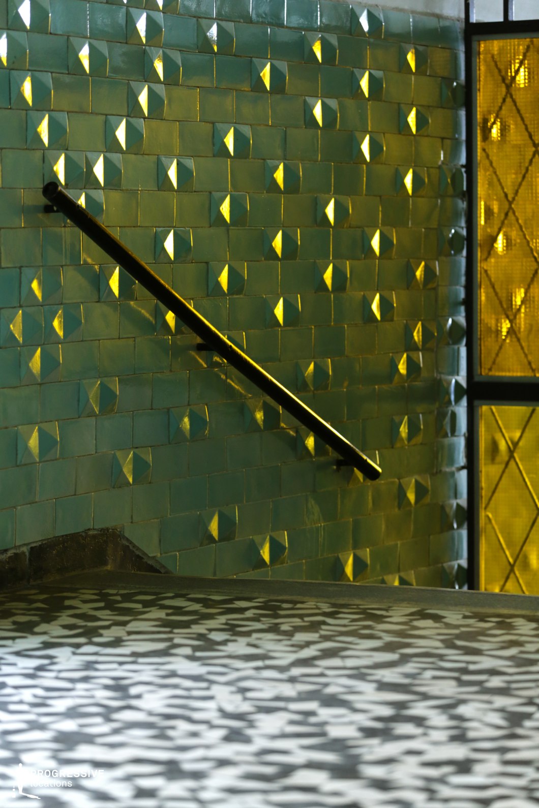 This photograph showcases a staircase with a wall covered in glossy blue tiles that have a unique, raised pyramidal texture. The tiles create an intricate pattern that reflects the surrounding light, producing a visually striking effect. A sleek black railing runs along the edge of the stairs, contrasting with the tiles. The light reflects off the tiles and the floor, creating abstract patterns that add dynamism to the space. On the right side of the image, a window with a yellow-tinted grid adds warmth to the cool tones of the tiles, contributing to the overall visual balance. This location, with its bold color contrasts and modern, geometric design, is perfect for films that require an industrial, artistic, or retro ambiance. Ideal for genres such as thrillers, mysteries, or science fiction, the space's distinctive style makes it well-suited for scenes set in futuristic or public environments like offices, subways, or unconventional stairwells.