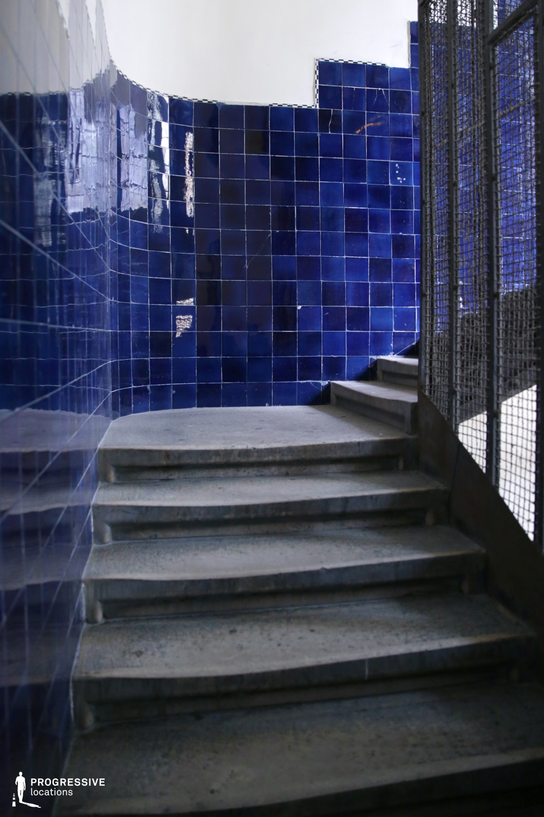 The image features a staircase surrounded by deep blue glazed tiles that line the curved walls. The tiles have a glossy finish, reflecting the light and creating a bold, almost reflective contrast with the rough, concrete steps. The metal railing on the right adds a raw, industrial element to the otherwise smooth and vibrant ceramic tiles. The space is minimalist yet striking due to the color contrast between the blue tiles and the neutral gray of the stairs. This location, with its modern industrial design, is ideal for film genres such as thrillers, crime dramas, or any film requiring a stark, visually engaging environment. Its bold color scheme and architectural features make it perfect for scenes set in urban stairwells, offices, or warehouses, adding both elegance and grit to a scene with its unique, high-contrast elements.