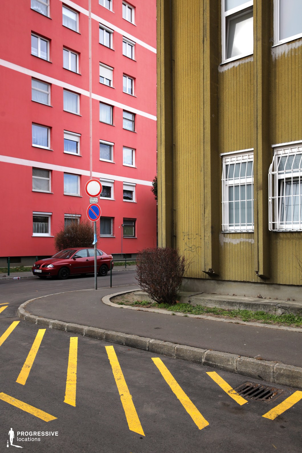 This image features a city street with a strong contrast between a yellow striped pavement and a mix of concrete buildings. The building on the left has a textured, yellowish-green facade, adding a retro, industrial feel, while the building on the right has a bold, red exterior with rows of windows. In the foreground, yellow diagonal lines on the asphalt emphasize the urban atmosphere, and there are signs and a parked red car nearby. This location, with its vibrant contrast and stark, functional design, works well for scenes requiring a gritty, urban backdrop. The combination of textured walls and yellow lines is perfect for modern thrillers, crime dramas, or dystopian films. Its industrial nature makes it an ideal setting for street-level scenes, parking lot interactions, or any urban environment that needs to feel both lived-in and somewhat utilitarian.