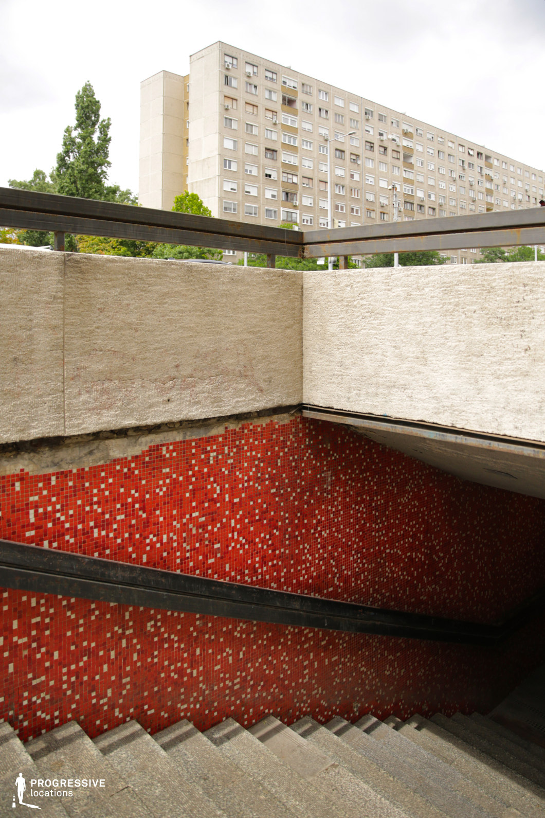 This photograph captures the interior of an underpass with a striking red mosaic tile wall that adds a vivid pop of color to the concrete space. The stairs, leading downward, are made of rough concrete with a simple black railing. The contrast between the bright red tiles and the neutral tones of the surrounding walls creates an industrial and somewhat retro atmosphere. The background reveals a large, functional residential building, enhancing the utilitarian feel of the space. This location is ideal for scenes in urban thrillers, crime dramas, or action sequences where the setting calls for a stark, gritty environment. The strong geometric patterns and bold color of the tiles make it suitable for film genres that require dramatic contrasts or urban settings. Its simple design can represent a variety of locations such as subway stations, underpasses, or public facilities, making it versatile for a range of narrative needs.