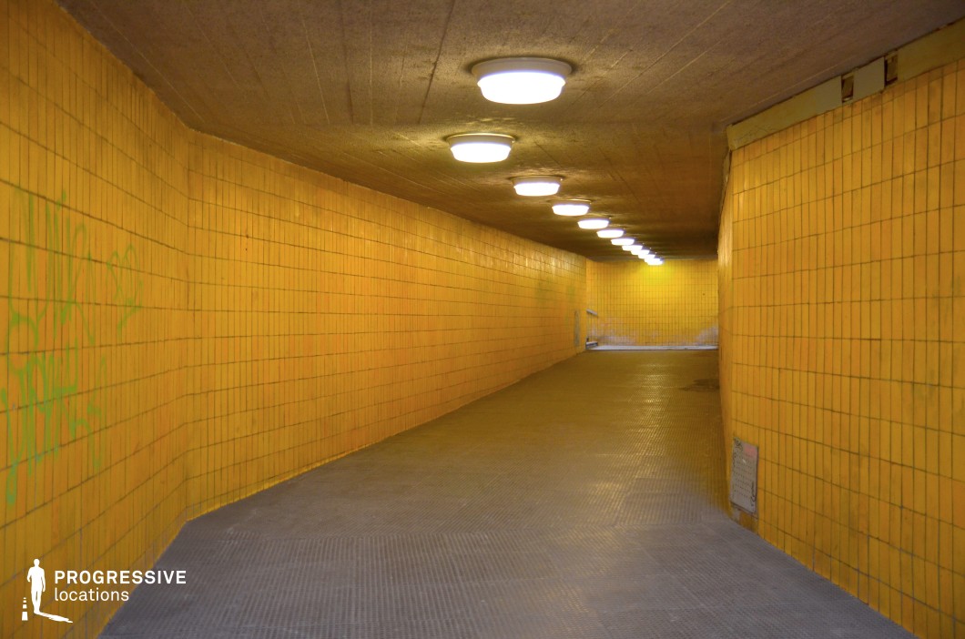 The image captures a bright yellow-tiled underpass, with the walls lined with square tiles that create a clean, yet industrial atmosphere. The tiles, though vibrant, show signs of wear, including some graffiti, which adds an urban edge to the scene. The lighting from the ceiling-mounted fluorescent lamps casts a uniform light across the corridor, enhancing the bold color of the walls. The textured floor further emphasizes the utilitarian nature of this space. This location is ideal for films that require a gritty, urban backdrop, such as crime dramas, thrillers, or action scenes. The stark yellow walls and industrial design evoke a sense of confinement, making it suitable for suspenseful or dramatic moments in scenes set in subway stations, pedestrian tunnels, or underground spaces.