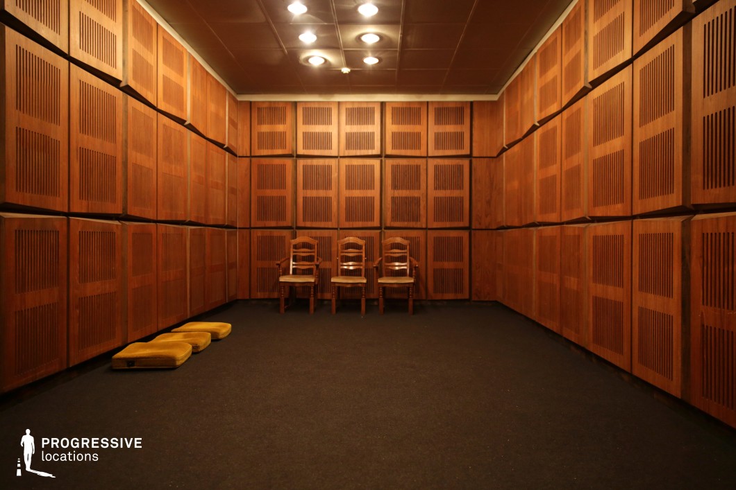 The photograph features a room with walls fully covered in wooden panels with vertical slats, creating a visually rich and symmetrical environment. The floor is covered with a dark carpet, and there are three wooden chairs placed against the wall, adding a sense of order and formality. The lighting is soft, with recessed ceiling lights providing even illumination across the space. This room exudes a warm, vintage atmosphere due to the rich wood tones and structured design. The simplicity and clean lines of the space make it versatile for a variety of film genres, particularly those that need a formal or slightly tense setting, such as dramas, thrillers, or psychological scenes. The warm, inviting wood texture, combined with the minimalist furniture, can also be ideal for scenes requiring a quiet, controlled environment, like interviews or intimate dialogues.