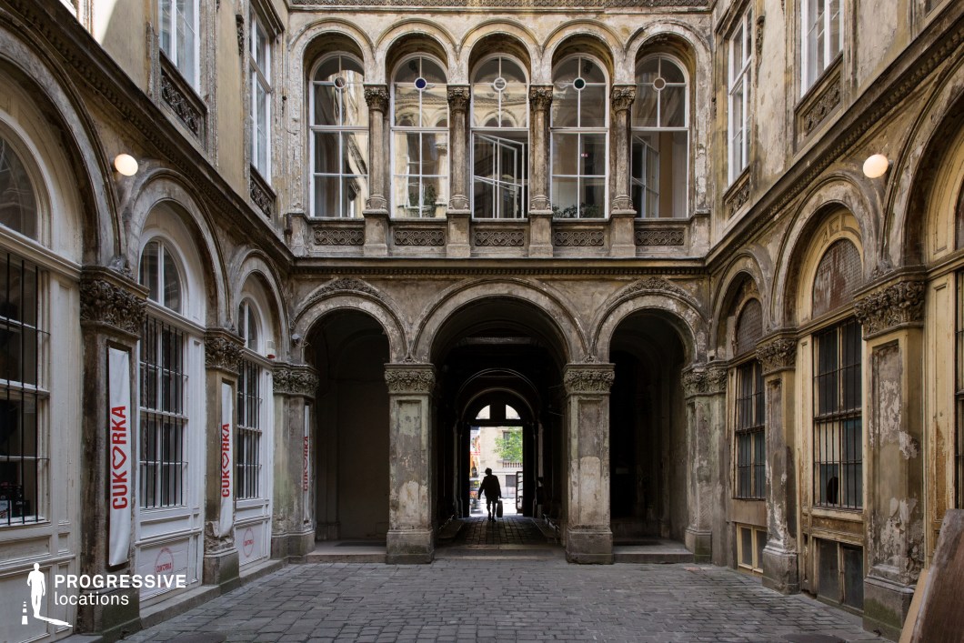 This richly detailed courtyard features classical architectural elements, including grand arches, decorative pilasters, and intricate stonework. The upper-level windows, framed by slender columns and adorned with delicate stained-glass accents, add an air of refinement. The aged patina on the walls, with peeling paint and weathered textures, enhances the historic character of the space. The central passageway, flanked by symmetrically arranged arches, leads to an external street, creating a strong visual depth. The blend of elegance and decay makes this an ideal filming location for period dramas, mystery thrillers, or historical narratives, offering a compelling mix of grandeur and timeworn beauty.