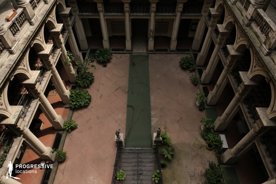 This image features a symmetrical courtyard enclosed by elegant two-story colonnades adorned with classical arches and ornate balustrades. The worn stone flooring, central pathway, and decorative stairway flanked by statues evoke a sense of faded grandeur. Lush greenery softens the structured architecture, adding an element of contrast. The weathered surfaces and aged patina enhance the location’s historical charm. This courtyard is ideal for filming period dramas, aristocratic intrigue, historical mysteries, or romantic narratives, providing a visually rich and atmospheric setting that blends elegance with a sense of nostalgia.