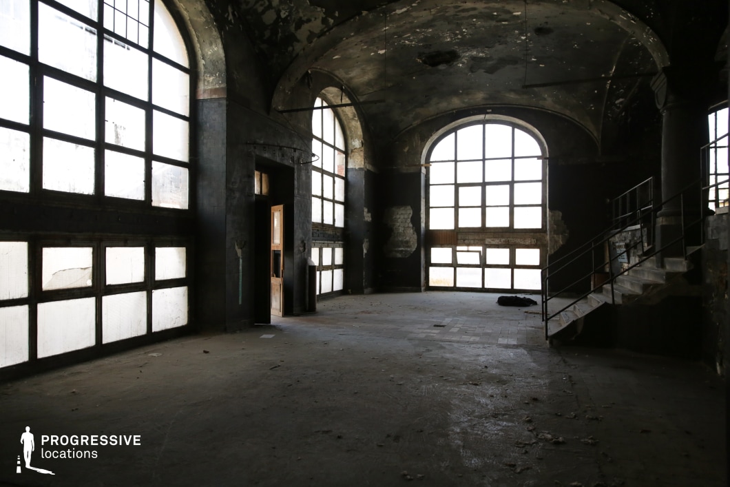 This image features a dark, abandoned industrial space with large arched windows, high ceilings, and decayed walls. The structure is dominated by blackened surfaces, peeling paint, and scattered debris, adding to the sense of desolation. The dramatic contrast between the dimly lit interior and the diffused light filtering through the multi-paned windows enhances the moody, cinematic atmosphere. A metal staircase and raised platform contribute to the industrial aesthetic. This location is ideal for filming dystopian narratives, crime thrillers, psychological horror, or post-apocalyptic settings, offering a raw and visually striking backdrop.