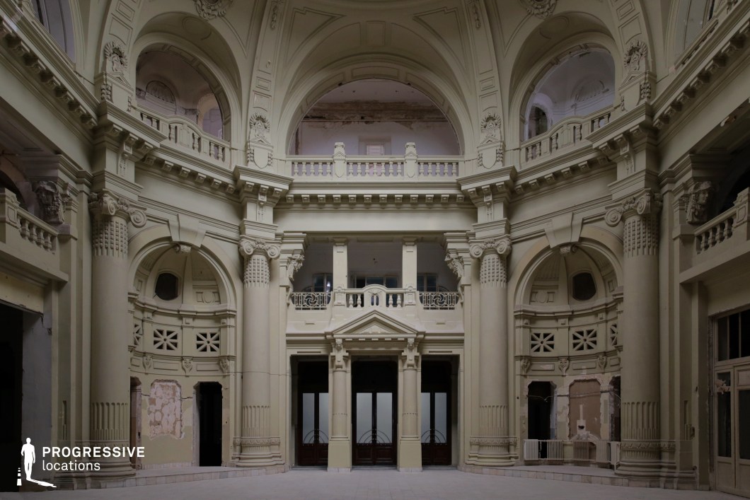 The image showcases a grand neoclassical interior with towering columns, intricate decorative moldings, and a series of elegant arches. The pale cream-colored walls and ornate detailing, including carved reliefs and balustrades, contribute to a regal and historic atmosphere. The hall's central focus is a grand entrance framed by massive pilasters and topped with a pediment, while balconies and upper walkways add a sense of scale and depth. Signs of wear and peeling plaster on the walls hint at an aged, abandoned, or partially restored state, enhancing the location's dramatic aesthetic. This space is ideal for filming historical dramas, period pieces, mystery thrillers, or even dystopian or fantasy settings, where opulence contrasts with decay to create a visually compelling backdrop.