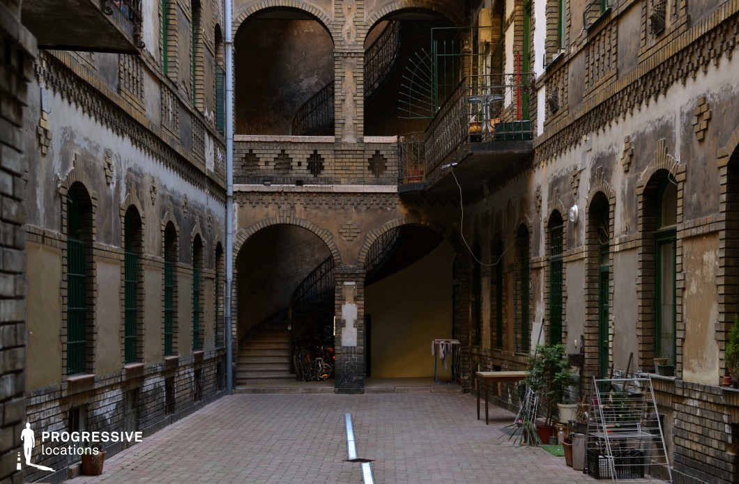 This image features a narrow, enclosed courtyard framed by weathered brick walls with repeating arched windows and doorways. The multi-level layout is accentuated by elegant curved staircases and small balconies, creating a dynamic sense of depth. The contrast between the faded plaster and exposed brick adds to the aged, atmospheric quality of the space. The worn surfaces and functional architectural details suggest a history of long-term habitation. This location is ideal for noir thrillers, urban dramas, period films, or crime narratives, offering a visually compelling and immersive setting that blends historical charm with a gritty, lived-in feel.