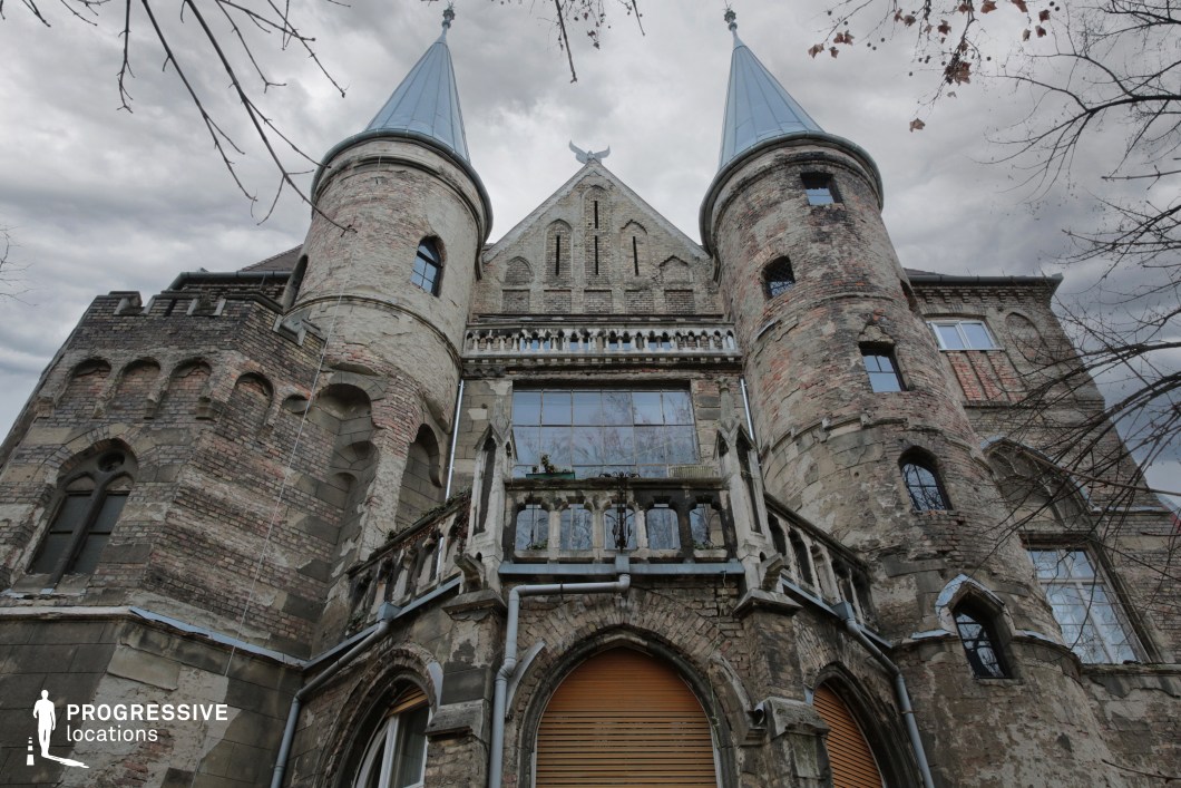 The image features an imposing, weathered castle with a Gothic architectural style, characterized by pointed turrets with metal roofs, arched windows, and intricate brickwork. The facade is heavily aged, with peeling plaster revealing underlying brick, cracked stone elements, and a partially collapsed balcony railing, enhancing its eerie, abandoned aesthetic. The upward perspective emphasizes the castle’s towering presence, while the intricate window designs and arched doorways contribute to its historical and mysterious character. This location is highly suitable for filming horror, fantasy, or gothic drama productions, offering an ideal backdrop for haunted mansion stories, supernatural mysteries, or medieval-inspired settings.