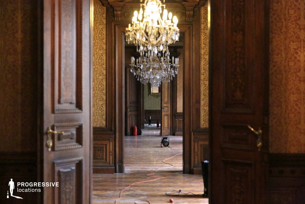 This image showcases a lavish palace corridor with a series of open wooden doors leading into progressively distant rooms, creating a strong sense of depth and symmetry. The warm-toned damask wallpaper, intricate wood paneling, and grand chandeliers contribute to the opulent atmosphere. The parquet flooring reflects the glow of the hanging crystal chandeliers, emphasizing the corridor's regal elegance. The blend of rich textures and classic architecture makes this an ideal location for filming period dramas, aristocratic intrigue, historical mysteries, or psychological thrillers, where a luxurious yet slightly aged setting enhances the narrative's depth.
