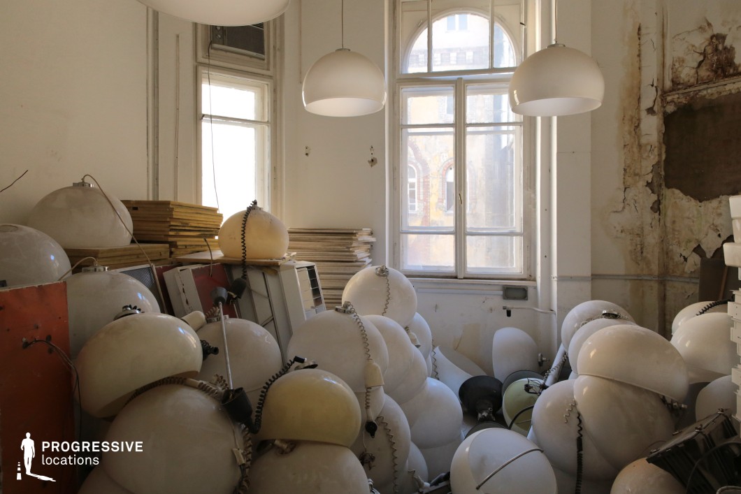 This dilapidated interior space features high ceilings, tall arched windows, and peeling white walls that reveal layers of age and neglect. The room is cluttered with a large pile of white dome-shaped lamps, some still attached to cords, creating a visually striking contrast between order and chaos. Hanging pendant lights cast a soft, muted glow, while the natural light filtering through the windows highlights the textured walls and aged surfaces. The classical window frames and the distant exterior view suggest a once-elegant building, now in disrepair. This location is ideal for filming surrealist dramas, psychological thrillers, or dystopian narratives, providing a unique and unsettling visual atmosphere that conveys themes of abandonment, memory, or artistic decay.