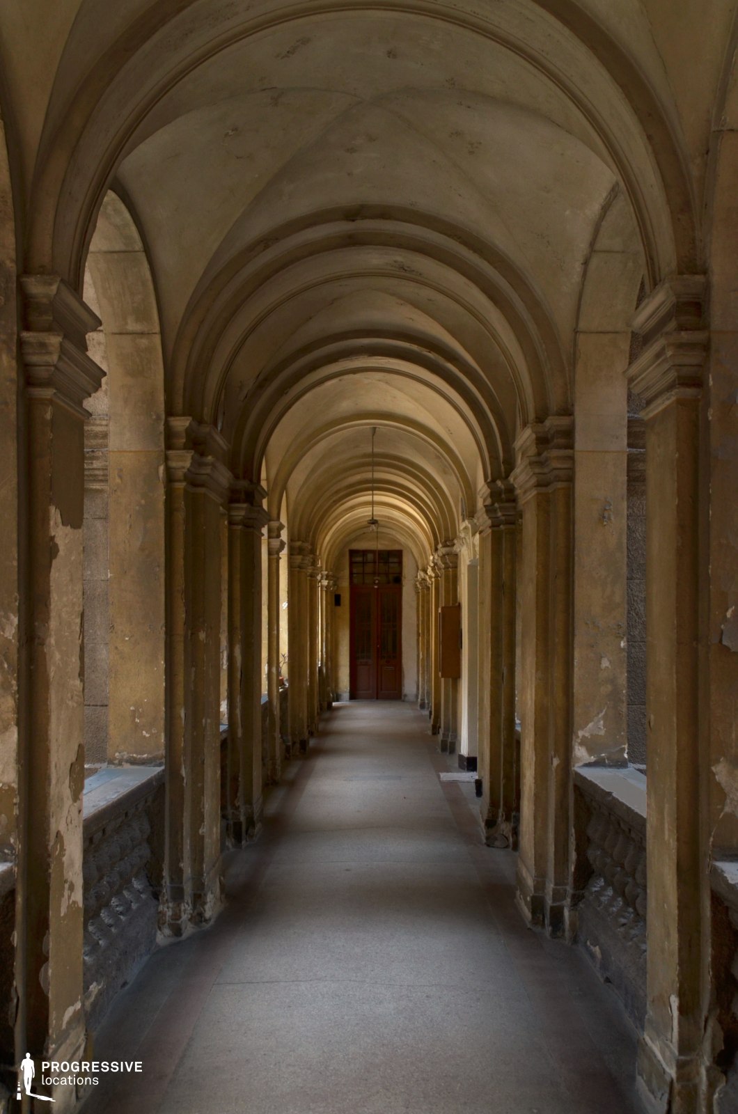 This long, arched corridor features a sequence of weathered stone columns supporting elegantly curved vaults, creating a rhythmic and immersive perspective. The walls show signs of age, with peeling plaster revealing the underlying structure, adding a sense of history and abandonment. The soft, diffused lighting enhances the depth of the passage, leading to a closed wooden door at the far end, which adds an element of mystery. The neutral, earthy tones of the architecture contribute to a timeless atmosphere. This location is ideal for filming historical dramas, suspense thrillers, or gothic-inspired narratives, offering a visually striking setting that conveys secrecy, intrigue, or the passage of time.