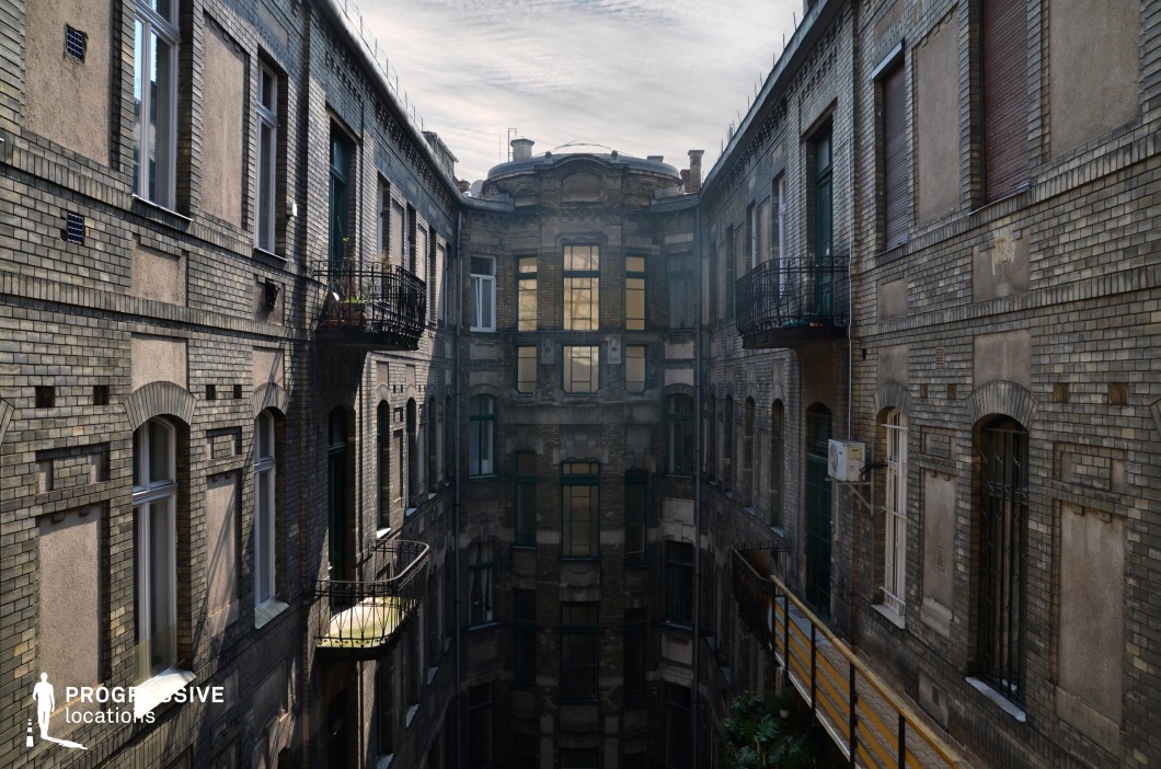 This enclosed courtyard is framed by tall, aged brick buildings featuring a mix of arched and rectangular windows, wrought-iron balconies, and intricate masonry details. The weathered facade, with patches of discoloration and peeling plaster, evokes a sense of history and decay. The perspective creates a strong sense of depth, drawing attention to the central, shadowed section of the structure, where multiple windows reflect light from an unseen source. The muted color palette of earthy browns, grays, and faded greens enhances the somber, slightly oppressive atmosphere. This location is ideal for filming suspenseful thrillers, noir dramas, or historical narratives, providing a visually rich backdrop that conveys tension, secrecy, and urban decay.
