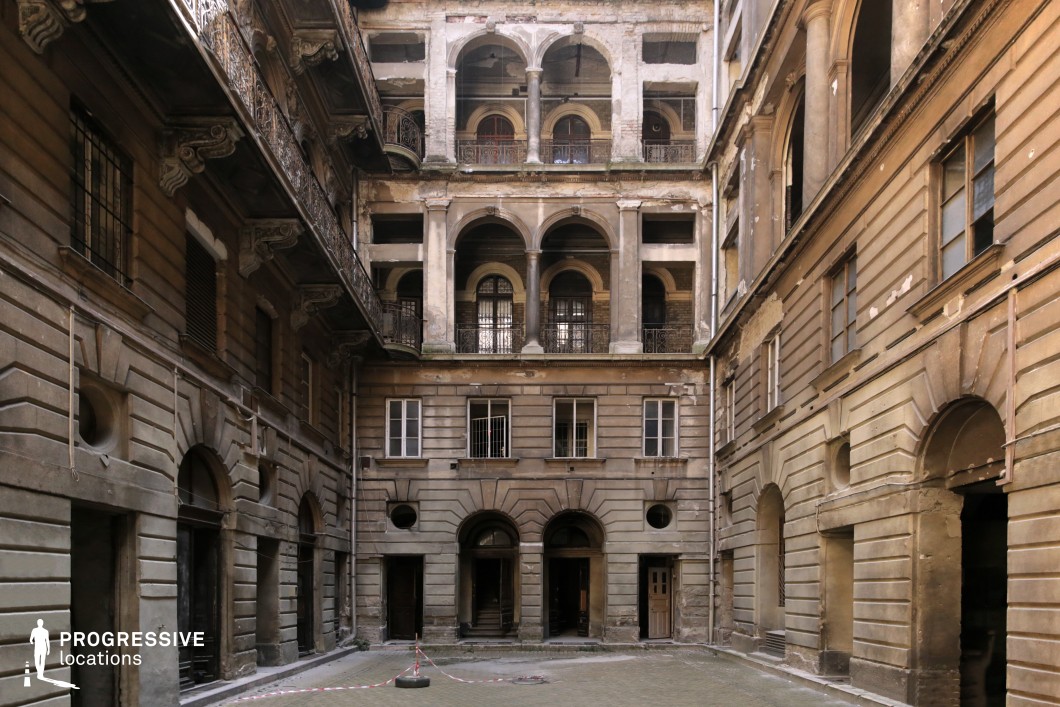 This grand yet deteriorating courtyard is enclosed by a multi-story building with classical architectural elements, including arched entryways, ornate balconies, and decorative stonework. The upper levels show signs of heavy wear, with crumbling plaster and exposed brick, contrasting with the symmetrical elegance of the structure. The warm, earthy tones of the aged facade, combined with deep shadows cast by the layered arches, create a striking visual depth. The space exudes a sense of forgotten grandeur, making it an ideal filming location for period dramas, historical thrillers, or noir-inspired narratives, offering a setting that conveys mystery, decay, and hidden stories.