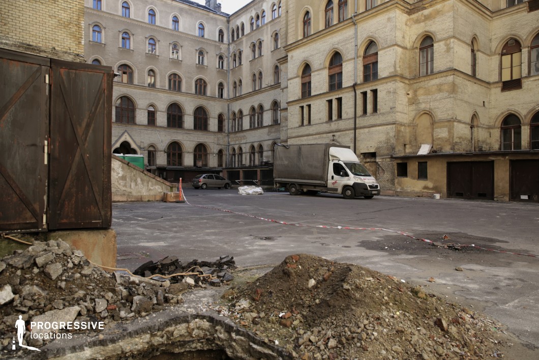 This enclosed courtyard is surrounded by a multi-story historic brick building with arched windows, faded facades, and visible signs of wear. The cracked asphalt ground, scattered debris, and construction remnants suggest partial neglect or ongoing restoration. Rusted metal doors and a weathered staircase contribute to the industrial, utilitarian atmosphere, contrasting with the grand architectural elements of the main structure. The muted color palette of beige and brown, combined with the rough textures, enhances the gritty, raw character of the space. This location is ideal for filming crime dramas, dystopian stories, or urban thrillers, offering a visually rich setting that conveys themes of abandonment, secrecy, and hidden histories.