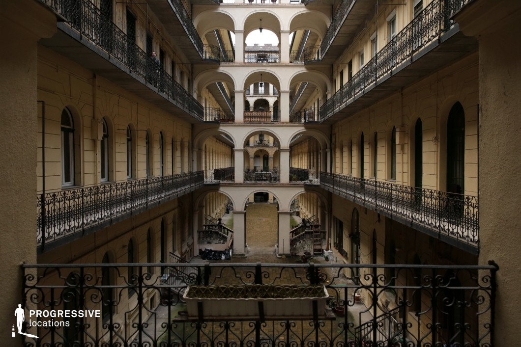 This image captures an expansive, multi-story courtyard enclosed by rows of evenly spaced arches and intricate wrought-iron railings. The symmetrical design, featuring a series of balconies overlooking a central open space, creates a striking visual rhythm. The warm-toned façade and classical architectural elements contrast with the aged surfaces, adding a sense of historic charm. The depth and perspective of the shot enhance the location’s cinematic appeal. This setting is ideal for filming period dramas, historical thrillers, urban intrigue, or noir-inspired narratives, offering a layered and atmospheric backdrop rich in architectural detail.