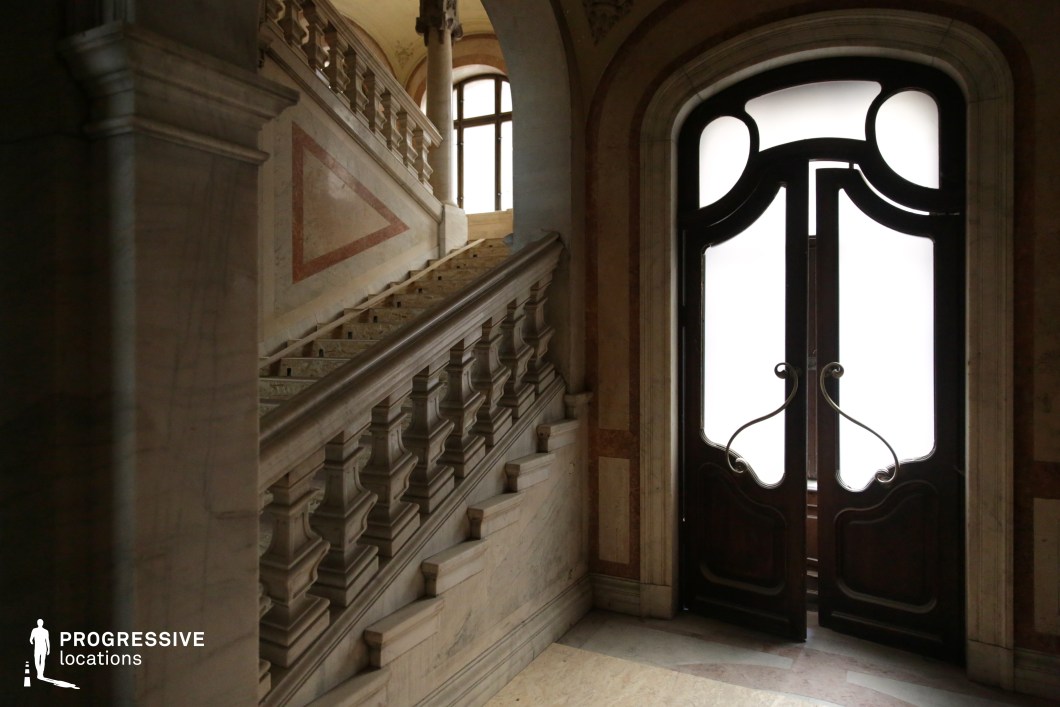 This grand interior features a sweeping marble staircase with intricately carved balustrades, leading to an upper level framed by classical columns. The polished stone surfaces, combined with decorative wall inlays and warm-hued accents, create a luxurious yet slightly aged atmosphere. The large double doors with curved wrought-iron handles and frosted glass panels add a sense of mystery and elegance. The architectural details suggest a historic European influence, making this location ideal for filming period dramas, aristocratic settings, or suspenseful thrillers where grandeur and intrigue play a central role.