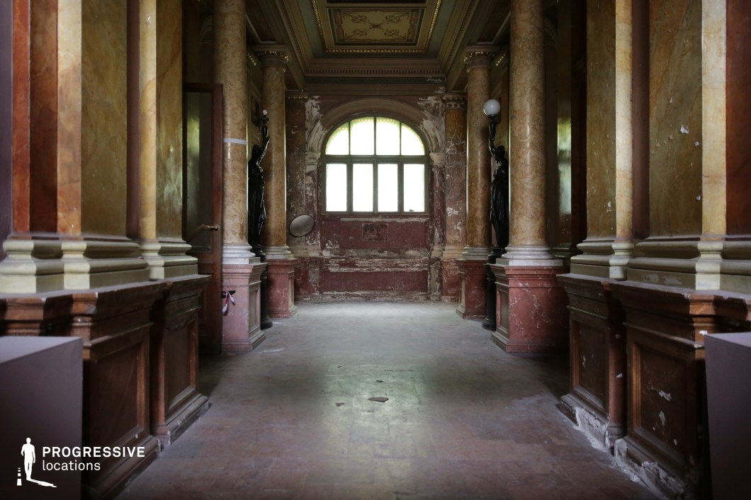 This image features an opulent yet decayed interior with richly veined marble columns lining a grand hallway. The warm tones of the polished stone contrast with the peeling paint and worn surfaces, emphasizing the building’s historic character. At the far end, a large arched window allows diffused light to illuminate the space, casting subtle reflections on the smooth floors. Decorative statues, integrated into the architecture, add a sense of grandeur and classical refinement. This location is ideal for filming historical dramas, aristocratic period pieces, supernatural thrillers, or mystery films, offering a striking blend of elegance and atmospheric decay.