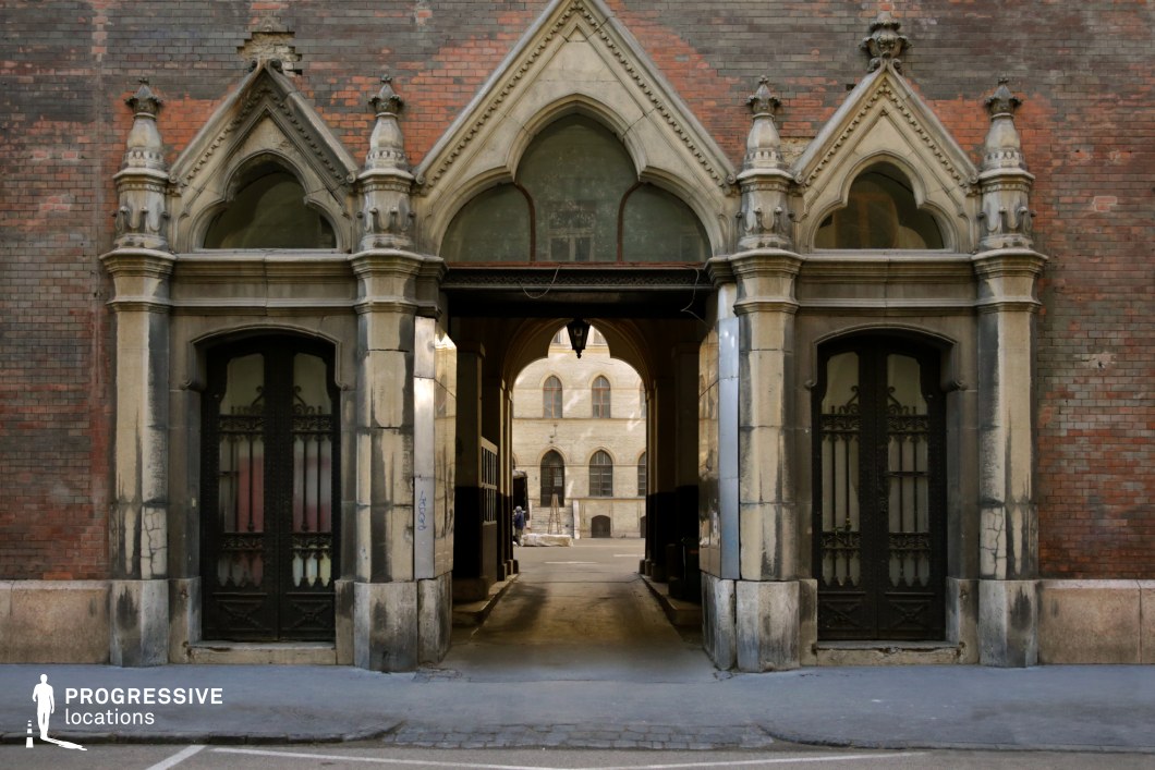 This image presents a grand, historic entranceway with pointed Gothic arches and decorative stonework. The facade, made of aged brick and weathered stone, showcases intricate architectural details, including ornamental finials and sculptural elements. Two large, wrought-iron doors flank the central arch, featuring elaborate patterns that add a sense of grandeur and secrecy. The passageway leads through a covered corridor, framed by further arches, into a courtyard with a classic European-style building in the background. This location is ideal for period dramas, mystery thrillers, or historical noir films, offering an atmospheric backdrop for intrigue, clandestine meetings, or dramatic arrivals.