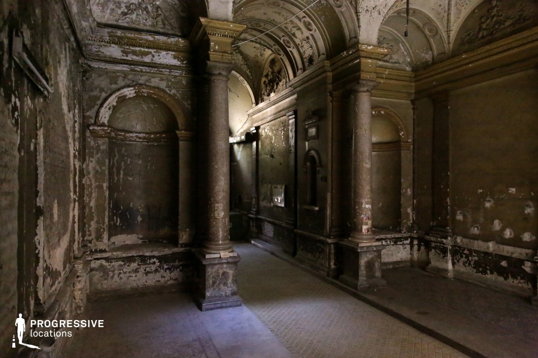 This image captures a dark, abandoned interior featuring ornate classical architecture with peeling walls, faded frescoes, and towering stone columns. The vaulted ceiling, adorned with intricate relief work, adds a sense of grandeur, while the chipped plaster and exposed surfaces emphasize the structure's age and neglect. Light filtering through distant openings creates dramatic contrasts between shadows and illuminated textures, enhancing the eerie and atmospheric quality of the space. This location is ideal for filming gothic horror, psychological thrillers, historical mysteries, or supernatural narratives, offering a haunting yet visually rich backdrop for dramatic storytelling.
