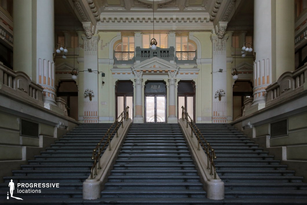 This image features an imposing neoclassical entrance hall with a wide, symmetrical staircase leading to a set of grand double doors framed by columns and ornate architectural detailing. The pale pastel walls, adorned with intricate moldings and sculptural reliefs, contrast with the darker stair treads and polished brass railings, emphasizing the space’s elegance. Decorative lighting fixtures and a high coffered ceiling enhance the historic atmosphere. This location is ideal for filming period dramas, aristocratic intrigue, mystery thrillers, or political dramas, offering a stately and commanding visual backdrop.