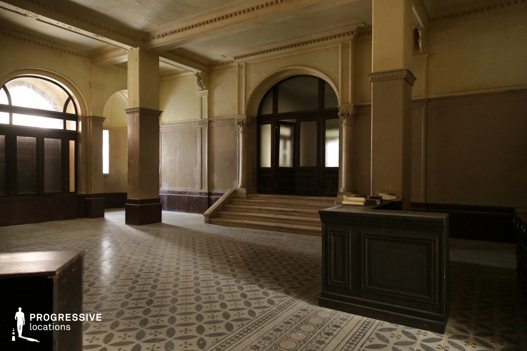This historic entrance hall features classical architectural elements, including tall, fluted columns with decorative capitals, arched doorways, and intricate crown molding. The patterned tile floor adds a refined, vintage aesthetic, while the worn surfaces and dim lighting evoke a sense of age and mystery. A grand staircase leads up to a set of wooden-framed glass doors, partially frosted, adding depth and intrigue. The muted color palette of beige, cream, and dark wood enhances the solemn yet elegant atmosphere. This location is ideal for filming period dramas, noir thrillers, or mystery narratives, offering a refined yet slightly eerie setting that complements themes of secrecy, authority, or historical significance.