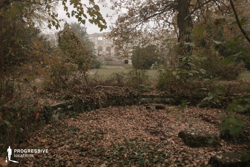 This atmospheric location features the remnants of a grand historic structure, now reduced to skeletal ruins in the distance, framed by dense foliage and an overgrown garden. The ground is blanketed with fallen leaves, while ivy and creeping vegetation reclaim stone structures, blending nature with the remnants of civilization. The muted earthy tones and soft natural light create a melancholic yet visually rich setting, evoking themes of decay and forgotten history. This location is ideal for filming historical dramas, fantasy narratives, or gothic mysteries, offering a dreamlike and hauntingly beautiful backdrop that conveys solitude, lost grandeur, and the passage of time.