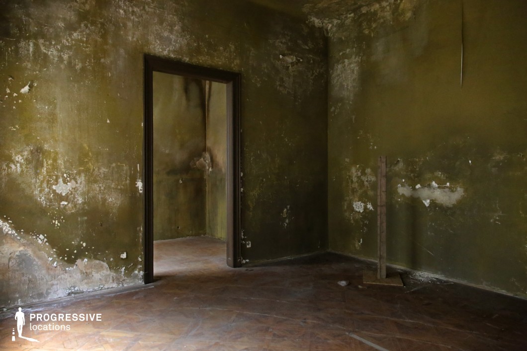 This abandoned interior features dark green-stained walls with peeling paint, cracks, and uneven discoloration, creating an eerie and unsettling atmosphere. The worn wooden floor, combined with the dim lighting and open doorway leading into another deteriorated space, enhances the feeling of desolation. The textured walls, marked by time and neglect, give the room a haunting quality. The muted, earthy tones and minimal furnishings add to its stark, almost cinematic emptiness. This location is ideal for filming psychological thrillers, horror films, or post-apocalyptic narratives, offering a setting that evokes decay, mystery, and psychological tension.