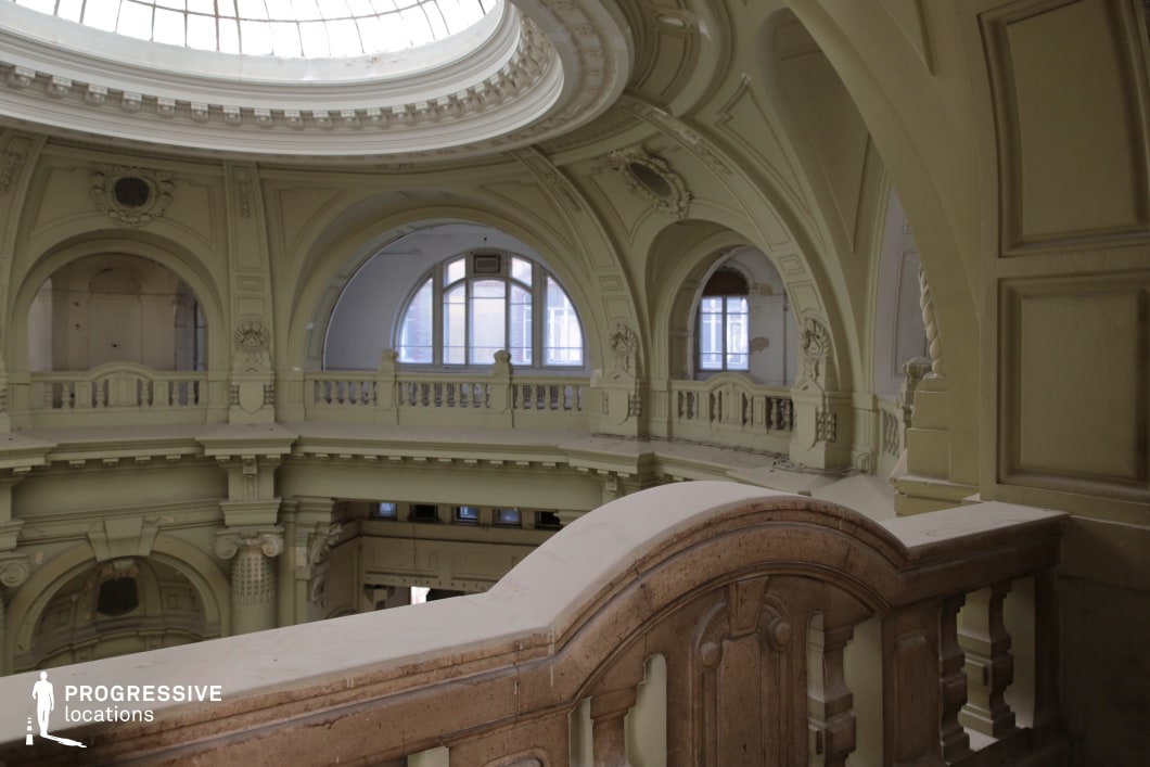 This image showcases an expansive, neoclassical interior featuring grand arched openings, intricate moldings, and a curved balcony overlooking the main hall. The pale-toned walls, adorned with sculptural reliefs and decorative balustrades, enhance the sense of elegance and architectural grandeur. A large, domed skylight floods the space with natural light, emphasizing the depth and symmetry of the structure. Subtle signs of wear add a layer of historical character. This location is ideal for period dramas, aristocratic narratives, mystery thrillers, or fantasy films, offering a sophisticated and visually striking setting that conveys both opulence and a sense of faded grandeur.